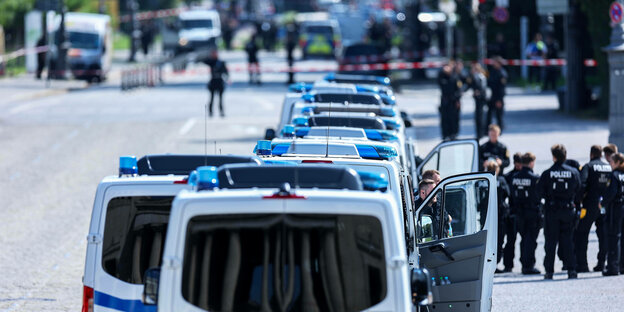 Mannschaftswagen der Polzie stehen in Reihe hintereinander, vorne stehen Polizisten in Uniform und im Hintergrund ein abgesperrter Tatort Mannschaftswagen der Polzie stehen in Reihe hintereinander, vorne stehen Polizisten in Uniform und im Hintergrund ein abgesperrter Tatort