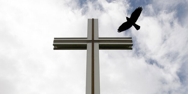Vor einem bewölkten Himmel steht ein Kreuz um den ein schwarzer Vogel fliegt Vor einem bewölkten Himmel steht ein Kreuz um den ein schwarzer Vogel fliegt