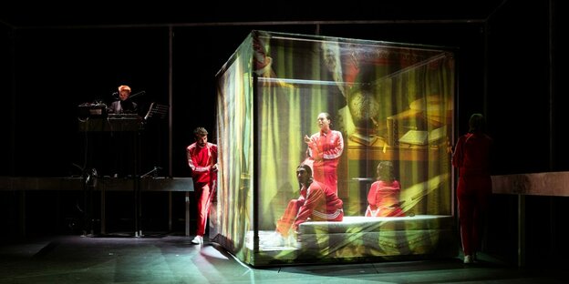 Menschen in roten Trainingsanzügen in einem Kubus auf einer Bühne Menschen in roten Trainingsanzügen in einem Kubus auf einer Bühne