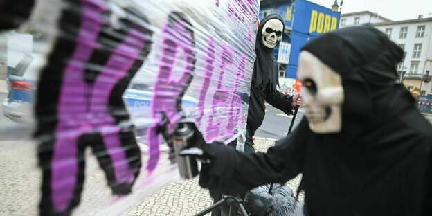 Ein Mensch mit einer Art Totenmaske sprüht in lila Buchstaben das Wort Krieg auf eine große Folie Ein Mensch mit einer Art Totenmaske sprüht in lila Buchstaben das Wort Krieg auf eine große Folie