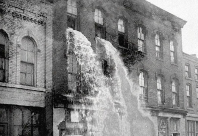 Historische Aufnahme von einem Gebäude in der Stadt, aus dessen oberer Etage Wasser in Strömen durch die Fenster nach außen fließt