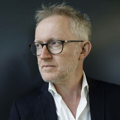 David Van Reybrouck, kurze graue Haare mit Scheitel, Brille, weißes Hemd, oberer Knopf offen und dunklen Blazer vor dunkelblauem Hintergrund im Profil von links fotografiert