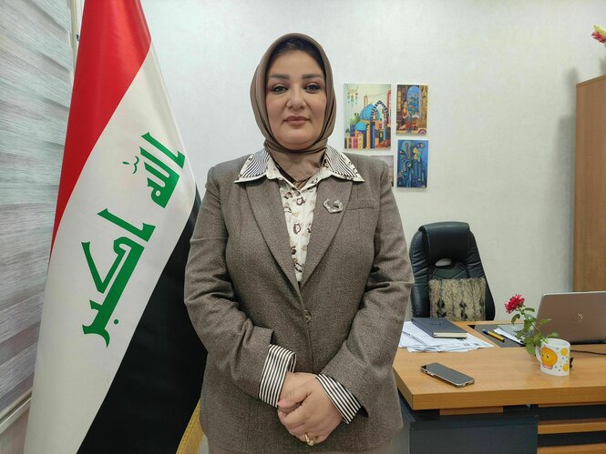 Nibras Abu Souda steht aufrecht in ihrem Büro vor einer irakischen Flagge, sie trägt ein Kopftuch, ein braunes Jacket und schaut ernst