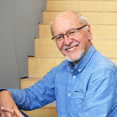 Armin Grundwald sitzt seitlich auf einer Treppe aus hellem Holz. Er trägt ein blaues Hemd, eine viereckige Brille, einen grau-weißen Bart und kurze, hellgraue Haare an den Seiten des Schädels. Er lächelt mit geöffneten Lippen in die Kamera.